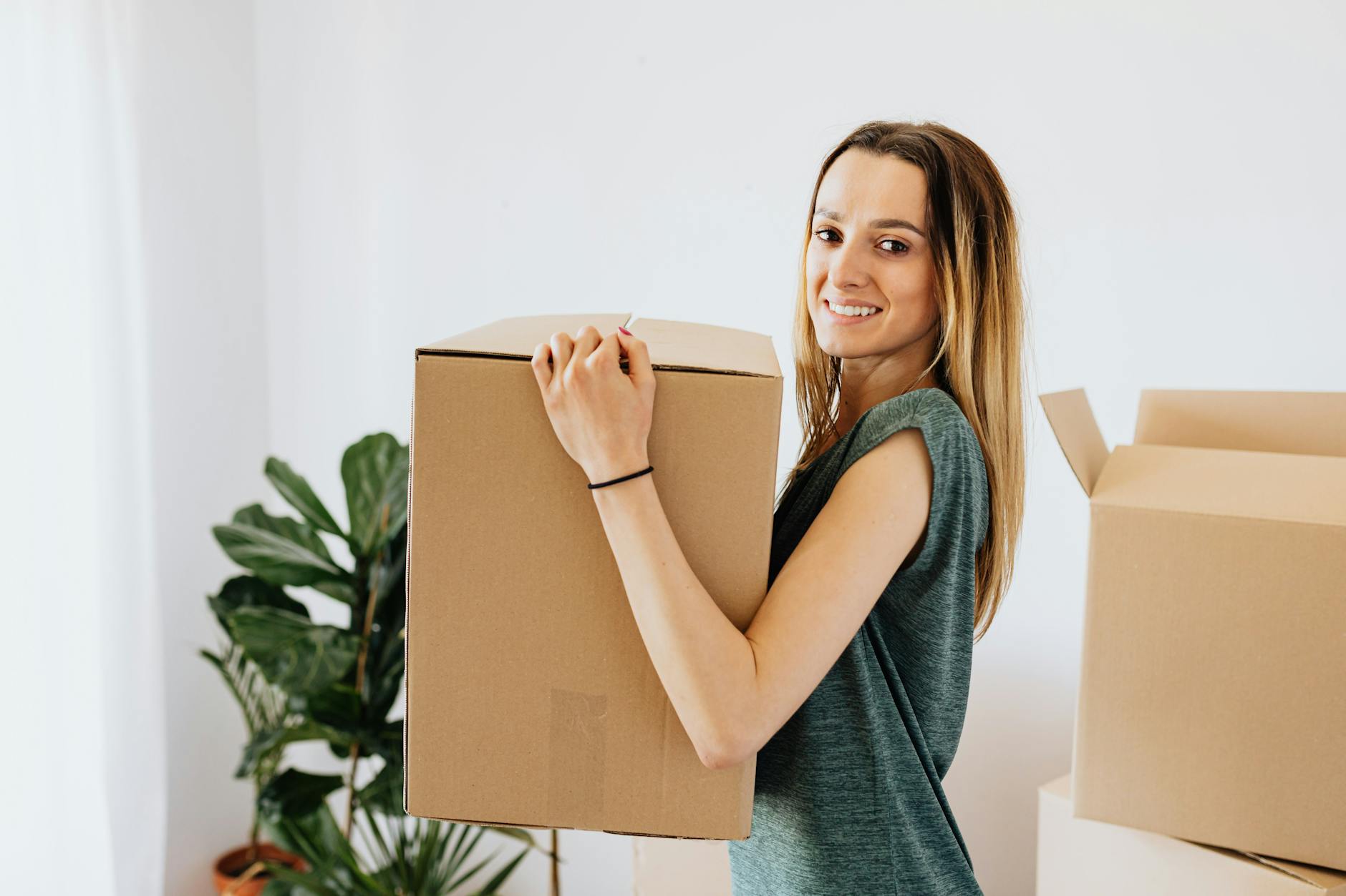 A smiling person carrying a cardboard box while moving into a new home