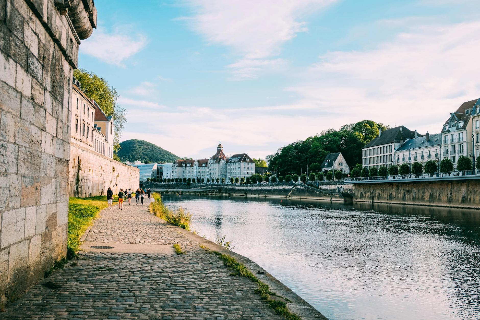 People strolling a riverside walkway past city architecture